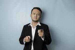 Portrait of young Asian businessman in black suit pointing index finger at camera, posing isolated over blue background. Cheerful smiling guy picking, choosing and indicating photo