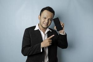 A portrait of a happy Asian businessman is smiling and holding his smartphone showing copy space on it's screen wearing black suit isolated by a blue background photo