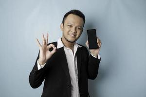 A portrait of a happy Asian businessman is smiling and holding his smartphone showing copy space on it's screen and gesturing Ok sign wearing black suit isolated by a blue background photo