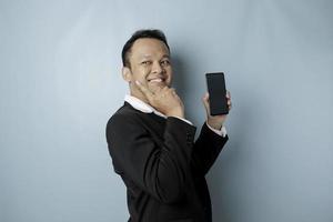 A portrait of a happy Asian businessman is smiling and holding his smartphone pointing copy space beside him wearing black suit isolated by a blue background photo