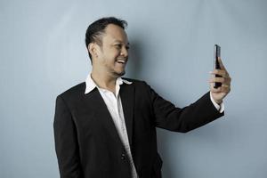 A portrait of a happy Asian businessman is smiling and holding his smartphone wearing black suit isolated by a blue background photo