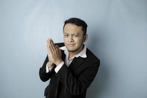 Smiling young Asian businessman wearing black suit gesturing greeting or namaste isolated over blue background photo