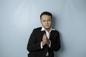 Smiling young Asian businessman wearing black suit gesturing greeting or namaste isolated over blue background photo