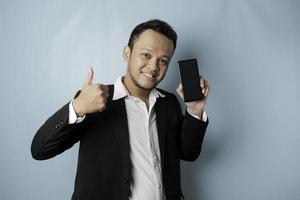 un retrato de un hombre de negocios asiático feliz sonríe y sostiene su teléfono inteligente que muestra espacio de copia en su pantalla con traje negro aislado por un fondo azul foto