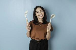 Excited Asian woman holding cooking ware and smiling, isolated by blue background photo