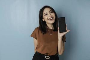 A portrait of a happy Asian woman is smiling and showing copy space on her smartphone wearing brown shirt isolated by a blue background photo