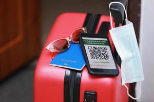 New normal concept. Close up view of a smartphone with an immune digital health passport, a red suitcase, protective mask, passport and sunglasses. Traveling by plane during a pandemic. photo