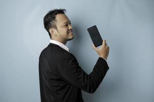 A portrait of a happy Asian businessman is smiling and holding his smartphone showing copy space on it's screen wearing black suit isolated by a blue background photo