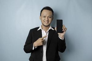 un retrato de un hombre de negocios asiático feliz sonríe y sostiene su teléfono inteligente que muestra espacio de copia en su pantalla con traje negro aislado por un fondo azul foto