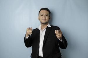 Portrait of young Asian businessman in black suit pointing index finger at camera, posing isolated over blue background. Cheerful smiling guy picking, choosing and indicating photo