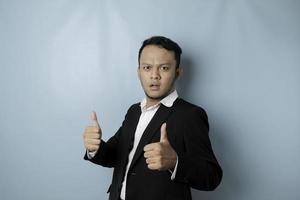 Excited Asian businessman wearing suit gives thumbs up hand gesture of approval, isolated by blue background photo