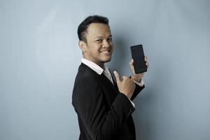 Excited Asian businessman wearing black suit gives thumbs up hand gesture of approval while showing copy space on his smartphone, isolated by blue background photo