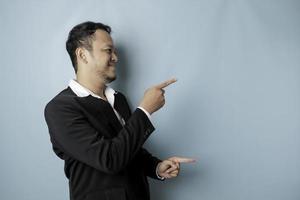 emocionado hombre asiático vestido con traje apuntando al espacio de la copia a su lado y sonriendo, aislado por fondo azul foto