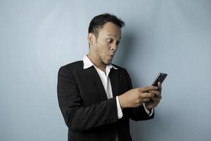 sorprendido hombre de negocios asiático con traje negro sosteniendo su teléfono inteligente, aislado por fondo azul foto