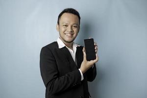 A portrait of a happy Asian businessman is smiling and holding his smartphone showing copy space on it's screen wearing black suit isolated by a blue background photo