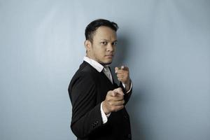 Portrait of young Asian businessman in black suit pointing index finger at camera, posing isolated over blue background. Cheerful smiling guy picking, choosing and indicating photo