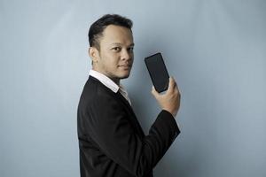 un retrato de un hombre de negocios asiático feliz sonríe y sostiene su teléfono inteligente que muestra espacio de copia en su pantalla con traje negro aislado por un fondo azul foto