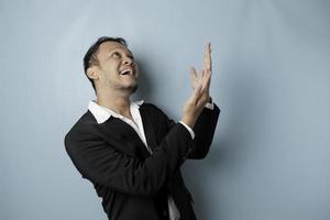 Excited Asian man wearing suit pointing at the copy space upside him, isolated by blue background photo