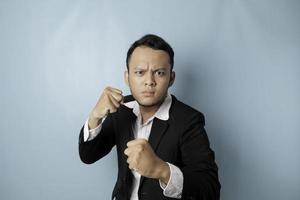 A portrait of a young Asian businessman looks angry and holding his fist ready to punch, isolated by blue background photo