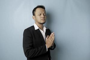 Smiling young Asian businessman wearing black suit gesturing greeting or namaste isolated over blue background photo