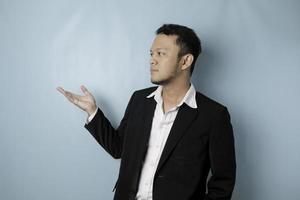 Excited Asian man wearing suit pointing at the copy space beside him and smiling, isolated by blue background photo