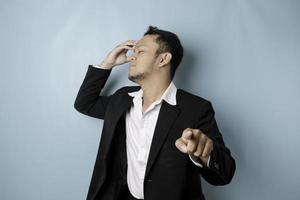 Portrait of young Asian businessman in black suit pointing index finger at camera, posing isolated over blue background. Cheerful smiling guy picking, choosing and indicating photo