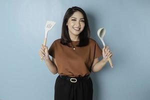 Excited Asian woman holding cooking ware and smiling, isolated by blue background photo