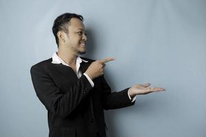 Excited Asian man wearing suit pointing at the copy space beside him and smiling, isolated by blue background photo