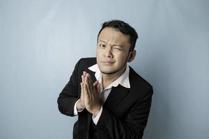 Smiling young Asian businessman wearing black suit gesturing greeting or namaste isolated over blue background photo