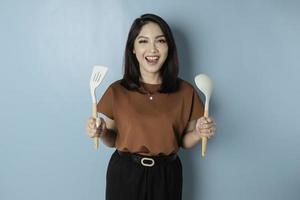 Excited Asian woman holding cooking ware and smiling, isolated by blue background photo