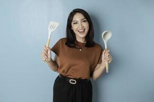 Excited Asian woman holding cooking ware and smiling, isolated by blue background photo