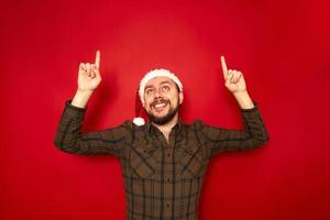 portrait of smiling man in Christmas hat casual clothes pointing his index fingers up at an empty space for advertising text isolated on red studio background concept people, recommendation, gestures photo