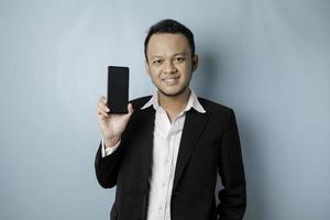 A portrait of a happy Asian businessman is smiling and holding his smartphone showing copy space on it's screen wearing black suit isolated by a blue background photo