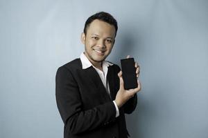 A portrait of a happy Asian businessman is smiling and holding his smartphone showing copy space on it's screen wearing black suit isolated by a blue background photo