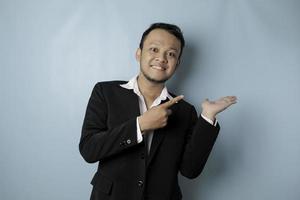 Excited Asian man wearing suit pointing at the copy space beside him and smiling, isolated by blue background photo