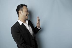 Excited Asian man wearing suit pointing at the copy space beside him and smiling, isolated by blue background photo