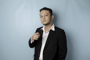 Portrait of young Asian businessman in black suit pointing index finger at camera, posing isolated over blue background. Cheerful smiling guy picking, choosing and indicating photo