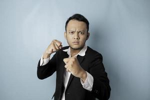 A portrait of a young Asian businessman looks angry and holding his fist ready to punch, isolated by blue background photo