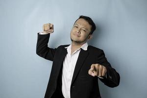 Portrait of young Asian businessman in black suit pointing index finger at camera, posing isolated over blue background. Cheerful smiling guy picking, choosing and indicating photo