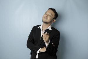 Portrait of young Asian businessman in black suit pointing index finger at camera, posing isolated over blue background. Cheerful smiling guy picking, choosing and indicating photo