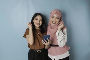 Two young Asian women are smiling while holding smartphone, isolated by blue background photo