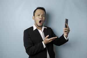 sorprendido hombre de negocios asiático con traje negro sosteniendo su teléfono inteligente, aislado por fondo azul foto