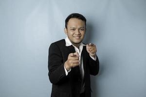 Portrait of young Asian businessman in black suit pointing index finger at camera, posing isolated over blue background. Cheerful smiling guy picking, choosing and indicating photo