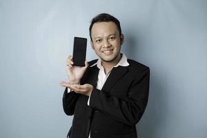 A portrait of a happy Asian businessman is smiling and holding his smartphone showing copy space on it's screen wearing black suit isolated by a blue background photo