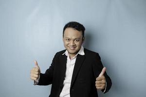 Excited Asian businessman wearing suit gives thumbs up hand gesture of approval, isolated by blue background photo