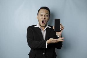 Surprised Asian businessman wearing a black suit holding his smartphone and showing copy space on it's screen, isolated by a blue background photo