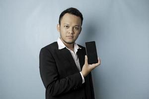 A portrait of a happy Asian businessman is smiling and holding his smartphone showing copy space on it's screen wearing black suit isolated by a blue background photo