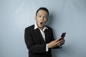 Surprised Asian businessman wearing black suit holding his smartphone, isolated by blue background photo