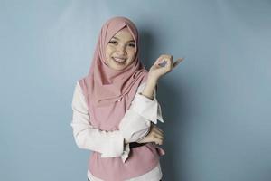 un retrato de una mujer musulmana asiática feliz sonríe y señala el espacio de copia a su lado, fondo azul aislado foto