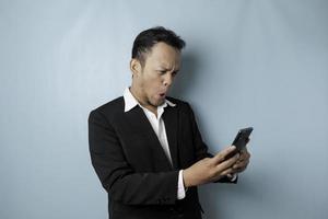 Surprised Asian businessman wearing black suit holding his smartphone, isolated by blue background photo